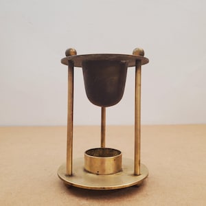 May include: A vintage brass candle holder. It features a circular base, a cylindrical cup, and three vertical supports. The candle holder has a small, round cup at the top. The design is simple and elegant, with a warm, metallic finish. The height is approximately 15 cm.