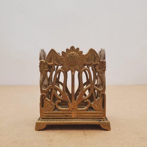 Peut inclure: Un organiseur de bureau en métal de couleur bronze avec un design Art nouveau. L'organisateur présente un motif de tournesol et floral, avec des motifs découpés complexes. La base rectangulaire a de petits pieds, et le design général est orné et décoratif.