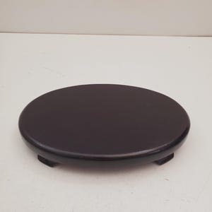 Op de afbeelding: Een ronde, donkerbruine houten displaystandaard met een glad, plat bovenoppervlak en een laag profiel. De standaard wordt ondersteund door kleine, geïntegreerde voetjes die de basis iets verhogen, wat een stabiel platform biedt voor het uitstallen van objecten.