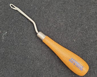 May include: A crochet hook with a wooden handle and a metal hook. The handle is light brown and has the text "Apostle Made in England" printed on it. The hook is silver and curved.