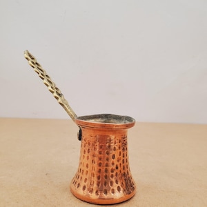 May include: A copper coffee pot with a brass handle. The pot has a textured surface with a flared base and a narrow neck. The handle is intricately designed with a repeating pattern.