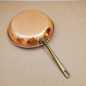 Könnte beinhalten: Eine Kupferpfanne mit Messinggriff. Die Pfanne hat eine runde Form und eine glänzende, reflektierende Oberfläche. Der Griff ist mit Nieten befestigt und hat ein Loch zum Aufhängen. Die Pfanne ist zum Kochen bestimmt.