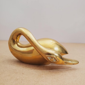 Pode incluir: Uma estatueta de latão polido em forma de cisne com um pescoço curvo e uma superfície lisa e reflexiva. O cisne é retratado em uma posição de repouso com a cabeça ligeiramente virada, exibindo um acabamento dourado metálico e forma aerodinâmica.