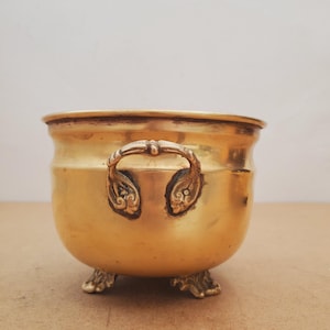 Puede incluir: Una pequeña maceta redonda de latón antiguo con asas ornamentadas y tres pies decorativos. La maceta tiene un acabado dorado pulido y un borde ligeramente ensanchado. Las asas están detalladas con volutas.