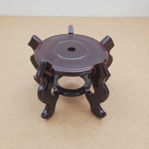 May include: A dark brown, circular display stand featuring a smooth, polished top surface with a central hole and four curved, ornate legs that extend outward. The stand is constructed with a sturdy base ring connecting the four legs for stability.