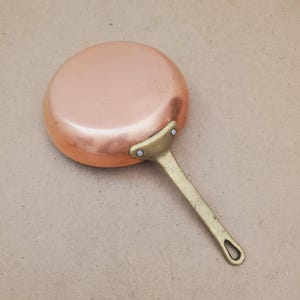 May include: A copper saucepan with a brass handle. The pan is round and has a polished surface. The handle is attached with rivets and has a loop for hanging. A classic piece of kitchenware, ideal for cooking.
