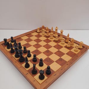 Può includere: Un set di scacchi in legno con una scacchiera a scacchi marrone chiaro e marrone scuro. I pezzi degli scacchi sono neri e marrone chiaro. La scacchiera è allestita per una partita a scacchi. Il set di scacchi è su una superficie bianca.