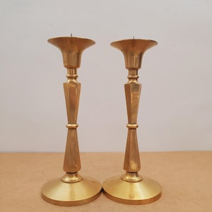 May include: Two gold-colored candlesticks with a classic design. Each candlestick has a wide, flared top, a faceted central column, and a round base. The candlesticks are made of metal and have a polished finish, suitable for home decor.