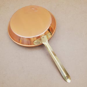 May include: A copper-colored frying pan with a long, brass-colored handle. The pan has a rounded shape and a shiny surface. The handle is attached to the pan with rivets. The pan is designed for cooking.