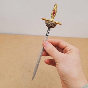 May include: Ornate letter opener with a gold-colored hilt and a silver blade. The hilt features a red jewel and intricate detailing. The letter opener is held in a hand against a neutral background.