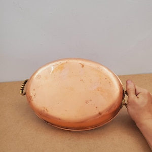 Op de afbeelding: Ovale koperen pan met messing handgrepen. De pan heeft een glanzend oppervlak met tekenen van slijtage. De handgrepen zijn gebogen en aan de zijkanten bevestigd. Dit vintage kookgerei is geschikt om in te koken en te serveren. Ongeveer 30 cm lang.