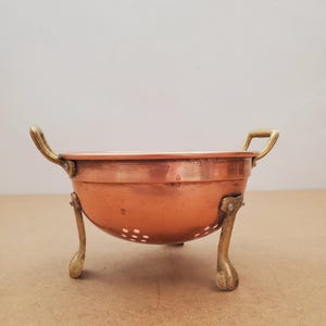 May include: A copper-colored bowl with two handles and three brass-colored legs. The bowl has a rounded shape with small holes on the sides. The handles and legs have a decorative design.