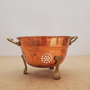 May include: A copper colander featuring a polished metallic finish, two small brass side handles, and three sturdy brass feet. The bowl is perforated with circular drainage holes arranged in a pattern around the lower section of the vessel.
