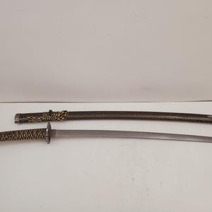 May include: A Japanese katana sword with a curved blade, resting beside its scabbard. The handle is wrapped in a textured, light brown and black pattern. The scabbard is dark with a gold-colored design.