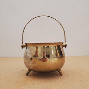 May include: A small, polished brass cauldron with a rounded body, three short legs, and a thin, arched metal handle attached to two side loops. The metallic surface shows signs of wear and oxidation, reflecting the surrounding environment.