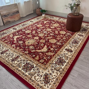 May include: A rectangular red area rug with an intricate floral pattern in beige and gold. The rug has a wide border with repeating floral and leaf motifs. The rug is placed on a light gray wooden floor.