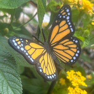 May include: A decorative butterfly with orange and black wings, detailed with black outlines and white spots, rests among green leaves and vibrant yellow flowers. The butterfly is a garden ornament.