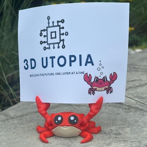 May include: A red crab figurine holding a white sign with the text "3D UTOPIA" and a circuit board graphic. The sign also features a cartoon crab illustration. The crab figurine is on a gray surface.