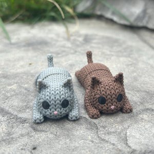 May include: Two small, knitted cat figurines. One is gray, the other is brown. Both have black eyes and pink noses. The cats are lying down on a stone surface. The knitted texture is visible.