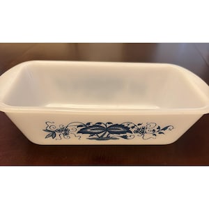May include: A rectangular white milk glass loaf pan featuring a blue floral and vine pattern printed on the exterior side. The baking dish has smooth, rounded edges and a deep, open interior basin designed for cooking or serving various food items.
