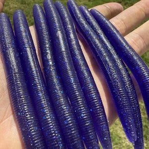 May include: A collection of dark blue, ribbed fishing lures with glitter accents. The soft plastic lures are cylindrical with a tapered end. The lures are held in a hand against a blurred green background.