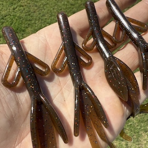 May include: A close-up of several brown fishing lures with glitter accents. The lures have a segmented body and multiple appendages, designed to mimic the appearance of prey. The lures are held in a hand against a background of green grass.
