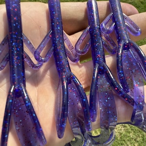 May include: Four purple fishing lures with red and blue glitter. Each lure has a segmented body, multiple appendages, and a curved tail. The lures are held in a hand against a background of green grass.