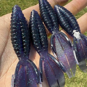 May include: Four dark purple fishing lures with a ribbed body and a split tail fin. The lures have a glittery, speckled appearance. The image is taken outdoors on a sunny day.