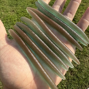 May include: A collection of soft plastic fishing lures in shades of brown and green, held in a hand against a backdrop of green grass. The lures have a ribbed texture and a tapered design, ideal for attracting fish.