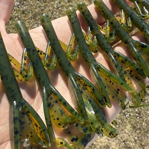 May include: A collection of green and yellow soft plastic fishing lures. The lures have a segmented body with a curly tail and are speckled with black dots. The lures are held in a hand against a blurred outdoor background.