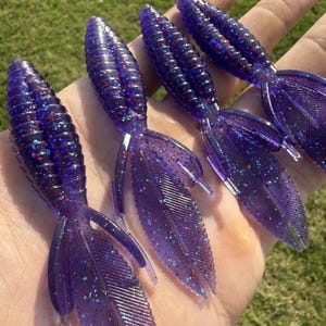 May include: Four purple fishing lures with glitter accents are displayed. Each lure has a ribbed body and a double-tail design. The lures are held in a hand against a backdrop of green grass, showcasing their vibrant color and detail.