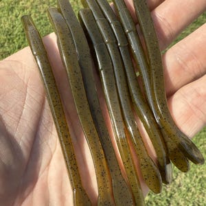 May include: A collection of eight olive-green soft plastic fishing lures. Each lure is slender, with a tapered body and a paddle tail. The lures are speckled with small black dots and are held in a hand against a background of green grass.