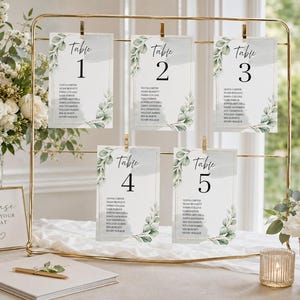 Könnte beinhalten: Ein goldener Metallständer mit fünf rechteckigen Sitzplan-Karten, beschriftet mit Table 1 bis 5, verziert mit grünen Aquarell-Eukalyptusblättern und Gästenamen. Ein kleines Schild trägt die Aufschrift please FIND YOUR SEAT neben einem Notizbuch.