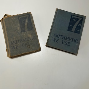 May include: Two vintage arithmetic textbooks. The books are a faded blue-gray color with the number 7 in a dark blue square on the cover. The text "ARITHMETIC WE USE" is printed below the number. One book shows signs of wear.