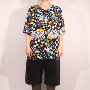 May include: A patterned button-down shirt in black, white, red, yellow, and blue geometric shapes. The shirt has short sleeves and a V-neck. Worn with black shorts.