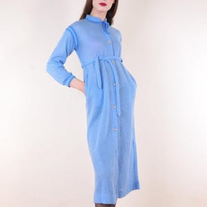 Puede incluir: Vestido azul claro de manga larga con un patrón texturizado. El vestido tiene botones en la parte delantera, un cinturón a juego y bolsillos. El vestido tiene un cuello alto y llega a la rodilla.