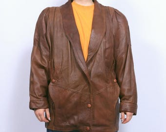 May include: A brown leather blazer with a notched collar and two front pockets. The blazer has a button closure and a tailored fit. The model is wearing a yellow t-shirt and blue jeans.