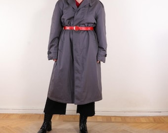 May include: A gray trench coat with a red belt. The coat is long, reaching the ankles, and has a classic design with a double-breasted front and epaulettes. The belt cinches the waist, creating a defined silhouette. The model is wearing black boots.