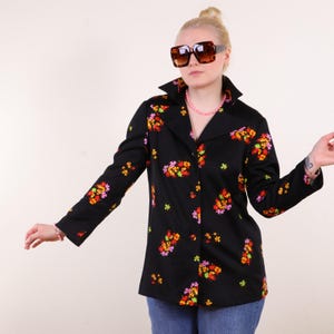 May include: A black blazer with a floral pattern in shades of red, orange, yellow, and pink. The blazer has a wide collar and is buttoned up. The person is wearing sunglasses and blue jeans.