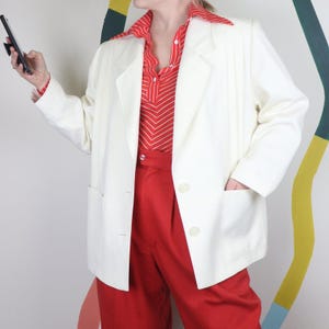 Puede incluir: Un blazer color crema con solapa de muesca y dos bolsillos delanteros. El blazer se lleva sobre una camisa a rayas rojas y blancas y pantalones rojos. El conjunto es de estilo vintage.