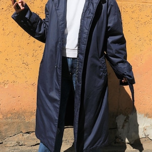 Peut inclure: Un long manteau bleu marine avec une fermeture boutonnée, un col à revers et des détails de ceinture aux manches. Le manteau est porté sur un haut blanc et un jean bleu. Le fond est un mur texturé orange-jaune.