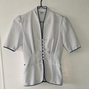May include: White blouse with a black polka dot pattern. The short-sleeved top has a V-neckline, a row of white buttons, and black trim along the edges. The blouse is hanging on a black hanger.