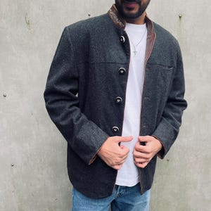 Op de afbeelding: Een donkergrijze jas met een bruine leren kraag en afwerking. De jas heeft een knoopsluiting en een opstaande kraag. Gedragen over een wit t-shirt en een blauwe jeans.