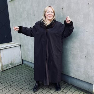 May include: A long, dark brown coat with a fur collar and cuffs. The coat is buttoned at the chest and reaches the ankles. The person is wearing black boots.