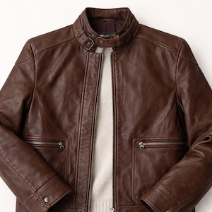 Puede incluir: Chaqueta de cuero marrón con cremallera, dos bolsillos con cremallera y cuello con botón a presión. La chaqueta tiene un diseño clásico con un acabado texturizado y un suéter de color neutro debajo.