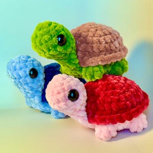 May include: Three handmade crochet turtles in shades of green, blue, pink, and red. Each turtle has a textured shell and black safety eyes. The turtles are stacked on top of each other, creating a playful and colorful display.