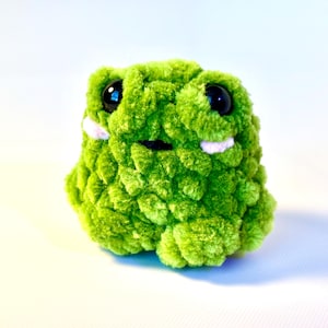 May include: A plush, green frog with large black eyes and small pink accents. The handmade stuffed animal has a textured, fuzzy appearance and a friendly expression, perfect for a child's toy or room decor.