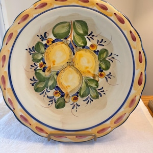 May include: A decorative ceramic plate with a scalloped edge. The plate features a hand-painted design of three yellow lemons with green leaves and blue accents. The rim has a yellow border with red and purple accents.