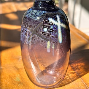 May include: A decorative glass vase with a dark blue rim and a body that fades from purple to clear. The vase features abstract white and brown designs. The vase is sitting on a wooden surface.