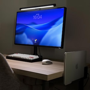Può includere: Un'impostazione informatica moderna con un monitor nero che mostra l'ora, una tastiera bianca con tasti rossi, un mouse bianco e un laptop argentato. Una barra luminosa a LED nera è montata sopra il monitor.
